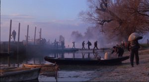Sur une lumière de crépuscule, des hommes portent des sacs sur les rives d'un petit fleuve sur lequel flottent des barques, scène du film Laurin.