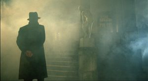 Un homme, de dos, avec un long manteau et un chapeau, patiente aux bas d'un escalier dans la brume, scène du film Laurin.