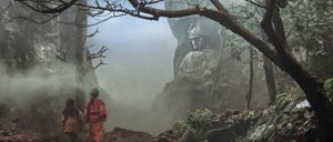 Plan d'ensemble large avec deux deux femmes dans une vallée brumeuse contemplant une immense statue de guerrier japonais taillée dans le flance de la montagne, plan d'un film de la trilogie Majin.