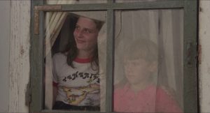 Deux enfants, une jeune fille et son petit frère, regardent à travers une vieille fenêtre brisée, d'un air narquois, scène du film Les révoltés de l'an 2000.