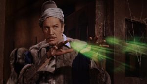 Vincent Price déguisé en mage lance un sort, des lumières vertes sortent de ses doigts dans le film Le corbeau.