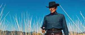 Gregory Peck en tenue de cow-boy sous un ciel bleu, dans un champ de blé, tient en joug un homme hors-champ avec son revolver dans le film Bravados.