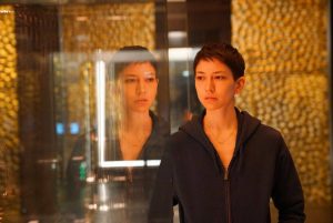 Lily Chan dans une pièce aux murs dorés, son visage se reflète dans une vitrine vide, scène de la mini-série Devs.