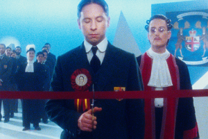 Le premier ministre s'apprête à couper le fil d'une inauguration, devant ce qui semble un juge et un groupe d'hommes en costume dans le film Le vingtième siècle.
