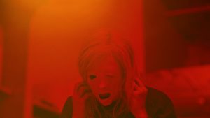 Sous une lumière rouge étrange, une femme porte les mains à ses oreilles, son visage est déformé comme s'il était en train de fondre, scène du film Possessor.
