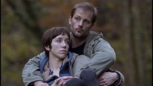 Dans une forêt, Sarah Giraudeau est assise devant Nicolas Duvauchelle qui la tient dans ses bras, tous deux regardent d'un air las dans une direction opposée, lui à gauche, elle à droite, scène du film Les envoûtés.