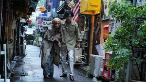 Mikura et Barbara marchent dans les rues de Tokyo, le dos voûtés, vêtus tous les deux d'un long pardessus gris.