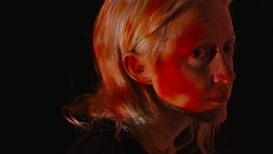 Plan rapproché-épaule du film Possessor où l'on voit Tanya Vos sur fond noir, une lumère rouge et blanche passe sur son visage