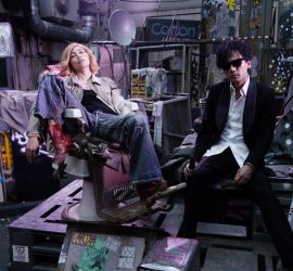 Le couple de héros de Tezuka's Barbara, lui en costume et lunettes noires sur le nez, elle en veste en jean et cheveux blonds décolorés, posent tous deux dans leur chambre-garage bordélique, regard caméra.