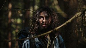 L'acteur Nat Wolff en homme des bois, cheveux hirsutes, longue  barbe, marche dans la forêt avec un sac sur le dos, scène du film Mortal.