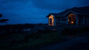 Une maison sur une falaise ua borde de l'océan de nuit, de la lumière est allumée sur le perron et dans le salon, mais il semble n'y avoir personne, plan du film The Rental.