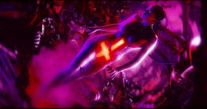 Une femme nue, une croix de lave sur le ventre avance dans une espèce de grotte indéfinie, dont les parois reflètent une intense lumière violette, scène du film Blood Machines.