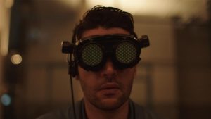 Gros plan sur un homme avec des lunettes futuristes, ressemblant à des yeux de mouche, dans le film Possessor.