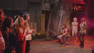 Un groupe d'hommes et de femmes souillés par des excréments font face à trois prostituées, dont l'une est en fauteuil roulant, scène du film Shakespeare's Shitstorm.