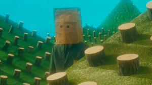 Un homme avec un sac en papier sur la tête est debout au milieu de colines miniatures sur lesquelles il y a de nombreux troncs d'arbres coupés, scène du film Le vingtième siècle.