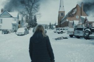 Aubrey vue de dos face à un petit village sous la neige, de la fumée noire flotte dans le ciel et s'échappe d'une maison sur la gauche qui vient de brûler, scène du Starfish.