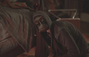 Agenouillé au pied d'un lit, un jeune homme à capuche tourne sa tête vers l'objectif, il porte un masque de singe en bois, scène du film I see you.