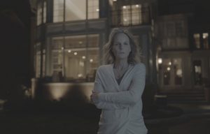 Helen Hunt devant son pavillon lumières allumées, croise les bras cheveux au vent, en regardant d'un air intrigue le trottoir d'en face, scène de nuit du film I see you.