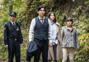 Dans une forêt, un homme en tenue de civil tient sa veste dans sa main, près de lui a sa droite sa femme et sa fille adolescente, tous les trois semblent inquiets ; derrière eux à gauche un agent de police, scène du film Inunaki.