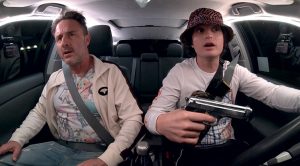Deux hommes dans une voiture : sur le siège conducteur un jeune homme porte un bob et tient un revolver dans sa main, sur le siège passager un homme d'âge mûr, scène du film Spree.