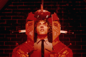 Sous une violente lumière rouge et devant un mur de briques, Tommy est inseré dans une armure métallique futuriste ; deux seringues sont plantées dans le métal au niveau des joues, plan du film Tommy.