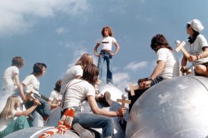 Tommy debout surplombe un groupe de jeunes, tous vêtus d'un t-shirt blanc et d'un jean bleu et asiss devant lui sur des boules d'acier.