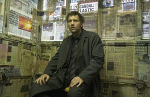 Clive Owen est assis dans une petite pièce aux murs sales et tapissés de pages de journaux, scène du film Les fils de l'homme.