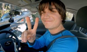 Joe Keery tout souriant au volant de son automobile, saluant en faisant un V avec ses doigts, dans le film Spree.