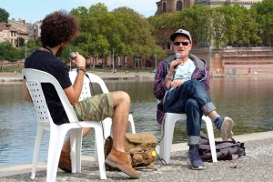 Au bord d'un cours d'eau à Toulouse, Jan Kounen est assis sur une chaise en plastique, face à son interviewer, un micro à la main.