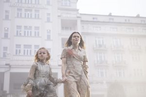 Une femme avec du sang sur l'épaule tient la main d'une petite fille ne lâchant pas son nounours, toutes les deux marchent dans une ville brumeuse qui semble déserte, scène du film Kadaver.