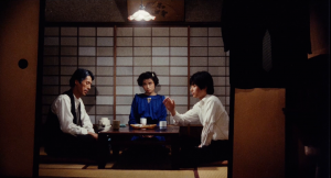 Dans un salon japonais, avec une unique ampoule au plafond diffusant une lumière blanche, dînent deux hommes en blanc et une femme, entre eux, vêtue en bleu.