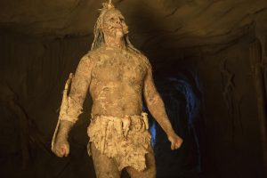 Un Amérindien du film Bone Tomahawk, colosse musclé recouvert de boue ocre, se tient debout prêt à frapper, dans une grotte.