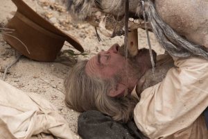 Kurt Russell allongé sur une terre aride, aux prises avec un bout de bois qu'on tente de lui enfoncer de force dans la bouche, scène du film Bone tomahawk.