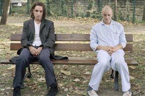 Sur un banc dans un parc défraichi, deux hommes sont assis ; celui de gauche en tenue de ville sombre est abattu, celui de droite semble sortir d'un asile de fous, chauve, vêtu d'un ensemble blanc, l'air inquiétant.