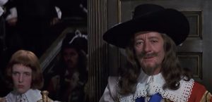 Scène du film Cromwell où ce dernier est debout contre une porte, regardant d'un oeil lmas devant lui, à sa gauche, dans des escaliers, des silhouettes du personnel de cour.