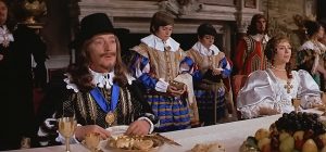 Charles 1er dîne à une longue table avec une nappe blanche, assis à sa gauche, sa femme la Reine, derrière eux leurs sbires, scène du film Cromwell de 1970.