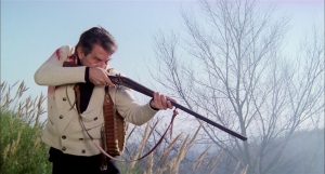 L'acteur Enrico Maria Salerno braque son fusil sur sa victime à terre, derrière lui un ciel brumeux et un arbre chétif dans le film La bête tue de sang froid.