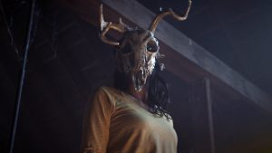 Une femme vue en contre-plongée, porte un angoissant masque représentant le crâne d'un cerf, scène du film The wretched.