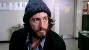 Al Pacino, barbe et bonnet, est assis dans une salle du commissariat, écoutant attentivement dans le fim Serpico.