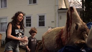 Une mère en short, t-shirt à tête de mort noir et sans manche, montre à son jeune fils la tête sectionnée d'une chèvre dans le film The wretched.