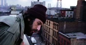 Serpico, alias Al Pacino en barbu et bonnet sur la tête est au sommet d'un immeuble new-yorkais, il se penche pour observer en bas, dans la rue.