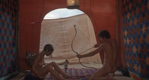 Dans une grande tente au milieu du désert et aux motifs or et bleus, un homme nu tend son arc vers l'entre-jambe d'une femme nue, assise face à lui, on devine une scène d'accouchement dans La trilogie de la vie de Pasolini.