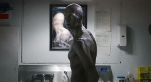 Un homme chauve et à la peau grise, squelettique, se tient debout, intrigué, dans un laboratoire, scène du film Depraved.