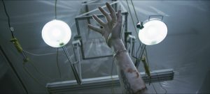 Un bras humain sectionné est relié à des fils et perfusions, entre deux agressives ampoules blanches dans le film Depraved.