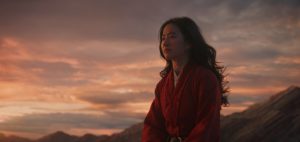 Mulan regarde l'horizon, derrière elle, un ciel de crépuscule orangé au dessus des montagnes.