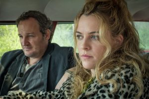 En voiture, Jason Clarke assis sur el siège passager, un rictus sur le visage, au volant Riley Keough plus sérieux, le regard vers l'horizon, portant un manteau léopard, scène du film Le diable tout le temps.