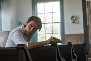 Robert Pattinson assis sur un des bancs en bois d'une église, se tourne vers derrière lui, jetant un regard sérieux, dans sa main droite, une Bible, scène du film Le diable tout le temps. 