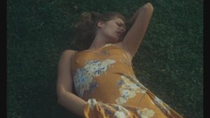 Une femme vêtue d'une robe jaune aux motifs floraux est allongée dans l'herbe, sa main droite passe sous sa robe, on devine qu'elle est en train de se masturber, scène du film Le jardin des supplices.