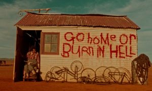 Au coeur du désert, une maison sur la quelle est taggé en rouge la phrase Go home or burn in hell, scène du film Spirits of the Air, Gremlins of the Clouds.