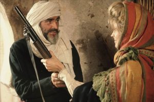 Une femme blonde donne un fusil à Sean Connery ; leurs regards à tous les deux sont étonanement doux ; tous deux sont vêtus de vêtements larges et d'un tissu leur couvrant le crâne comme de coutume dans le désert ; scène du film Le lion et le vent.
